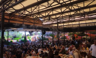 A high-angle shot of a large, crowded outdoor garden restaurant with a metal-framed canopy, misting fans, and hundreds of people dining at tables, set against a backdrop of vibrant, multicolored flower hills and rock waterfalls.