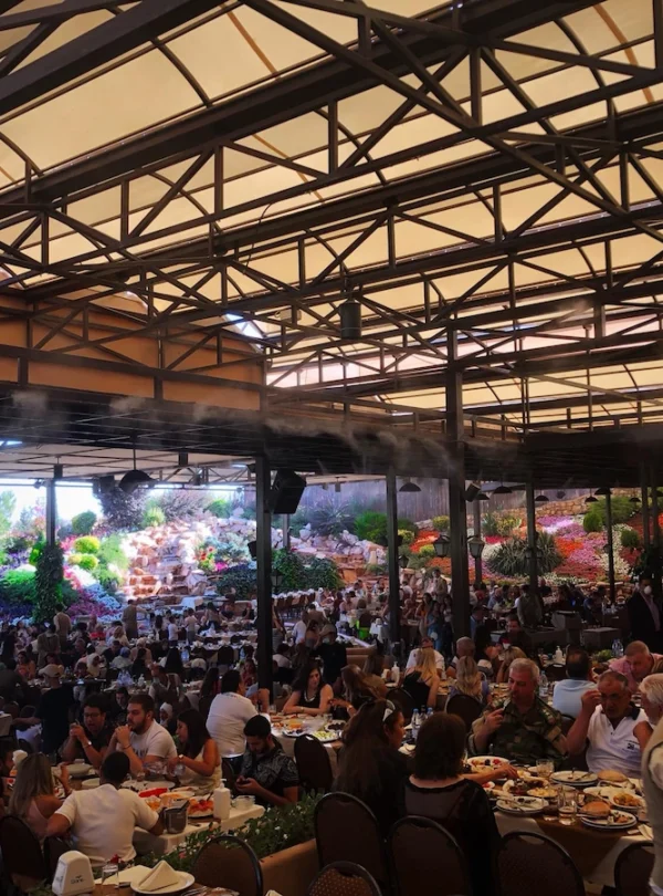 A high-angle shot of a large, crowded outdoor garden restaurant with a metal-framed canopy, misting fans, and hundreds of people dining at tables, set against a backdrop of vibrant, multicolored flower hills and rock waterfalls.