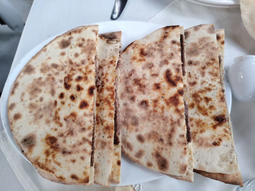 A close-up view of a white plate holding four large, semi-circular pieces of toasted Syrian flatbread stuffed with a thin layer of seasoned minced meat, showing golden-brown grill marks on the surface.