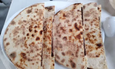 A close-up view of a white plate holding four large, semi-circular pieces of toasted Syrian flatbread stuffed with a thin layer of seasoned minced meat, showing golden-brown grill marks on the surface.