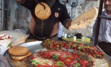 A street food vendor holds two large, thin, disc-shaped pieces of Kibbeh Sajieh that have been deep-fried to a golden brown, standing over a large circular tray of sautéed red and green peppers.