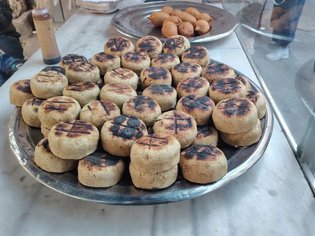 A large silver platter piled high with round, thick patties of grilled kibbeh featuring dark char marks from the fire, displayed on a marble counter at a local Syrian food stall.
