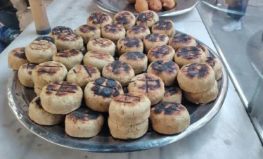 A large silver platter piled high with round, thick patties of grilled kibbeh featuring dark char marks from the fire, displayed on a marble counter at a local Syrian food stall.