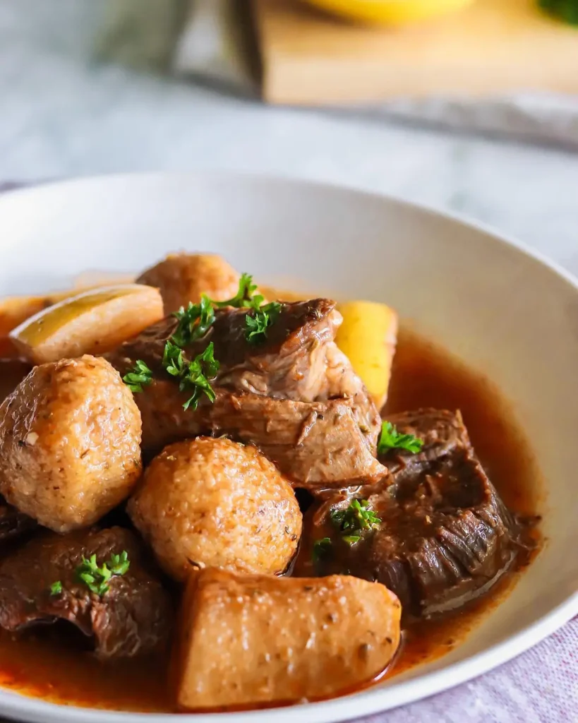 A close-up, top-down view of a white ceramic bowl containing Kibbeh Esfarjelieh, featuring golden-brown fried kibbeh balls simmered in a rich, reddish-brown sauce with chunks of cooked quince and tender meat, garnished with fresh green parsley.