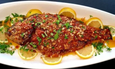 A close-up, top-down view of a whole baked fish served on a white platter, generously topped with a bold, red spicy sauce made from peppers, tomatoes, and walnuts, garnished with fresh parsley and lemon slices.