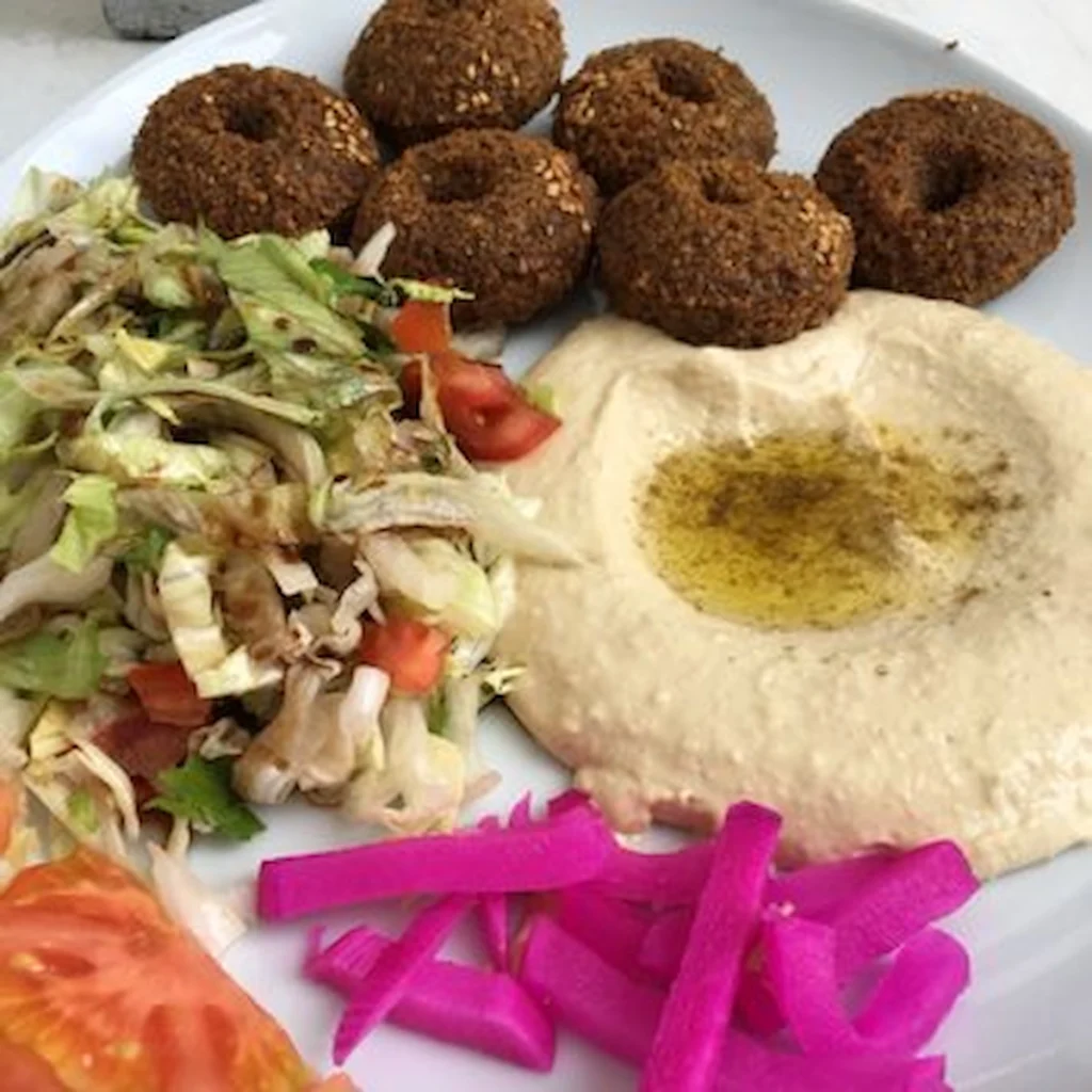 A white plate featuring five crispy, donut-shaped falafel pieces with sesame seeds, a large swirl of creamy hummus topped with olive oil, a fresh vegetable salad, and bright pink pickled turnip strips.