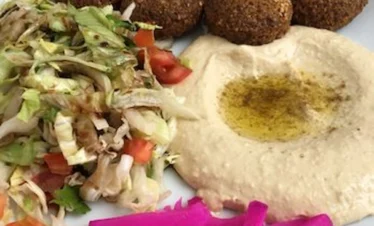 A white plate featuring five crispy, donut-shaped falafel pieces with sesame seeds, a large swirl of creamy hummus topped with olive oil, a fresh vegetable salad, and bright pink pickled turnip strips.