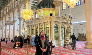 A female traveler wearing a traditional black and gold abaya stands in the vast, carpeted interior of the Umayyad Mosque in Damascus, with ornate chandeliers, white marble columns, and a green-domed shrine in the background.