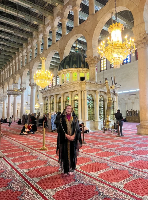 A female traveler wearing a traditional black and gold abaya stands in the vast, carpeted interior of the Umayyad Mosque in Damascus, with ornate chandeliers, white marble columns, and a green-domed shrine in the background.