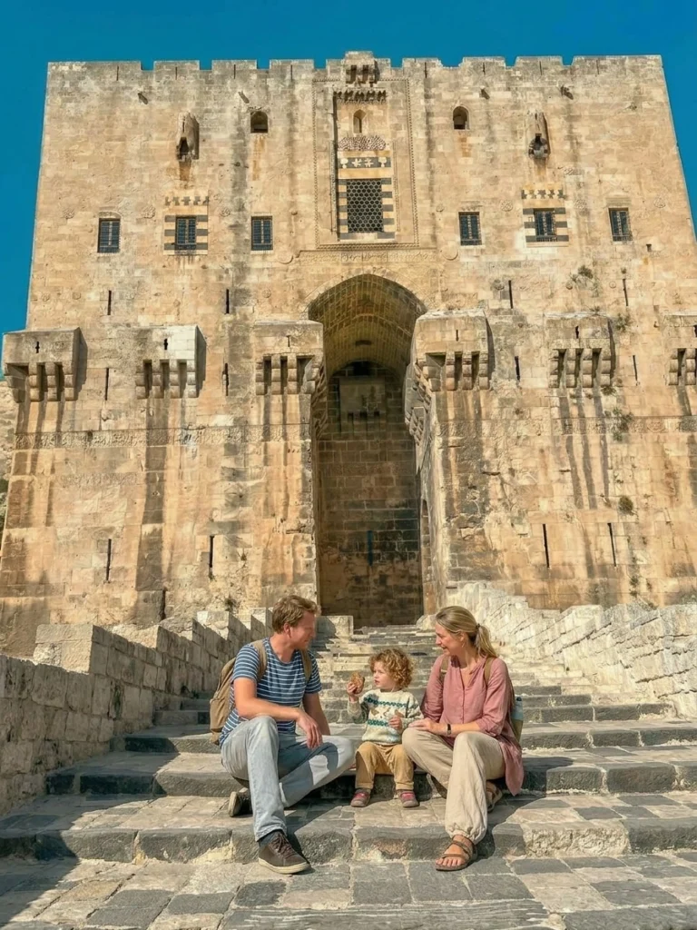 A candid, sun-drenched photograph of a family of three (a man, woman, and young child) sitting together on the large stone steps leading up to the colossal main gateway of the Citadel of Aleppo