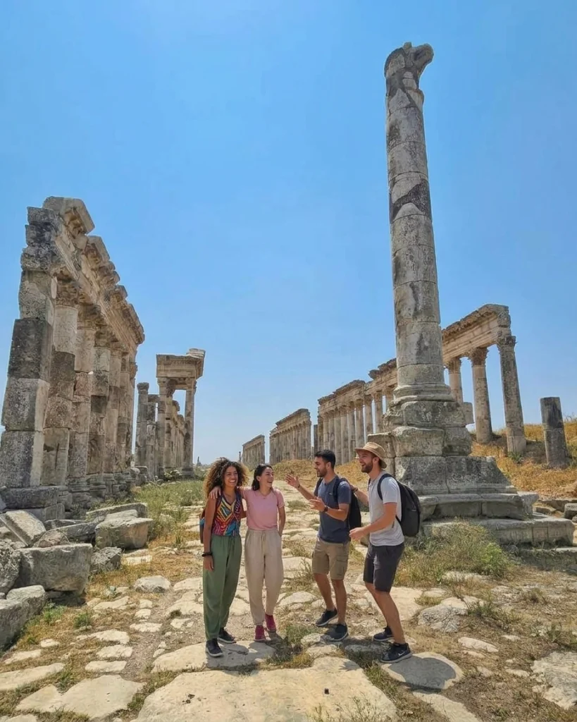 A wide-angle, sun-drenched photograph of a group of four diverse young tourists (two women and two men) in modern travel clothing, laughing and walking along the ancient, weathered stone-paved Roman street in Apamea, Syria[cite: 19]. The perspective looks down a massive, iconic avenue flanked by rows of monumental columns, with the large, detailed broken column section featuring a lion motif prominent in the right foreground against a clear blue sky[cite: 19, 20].