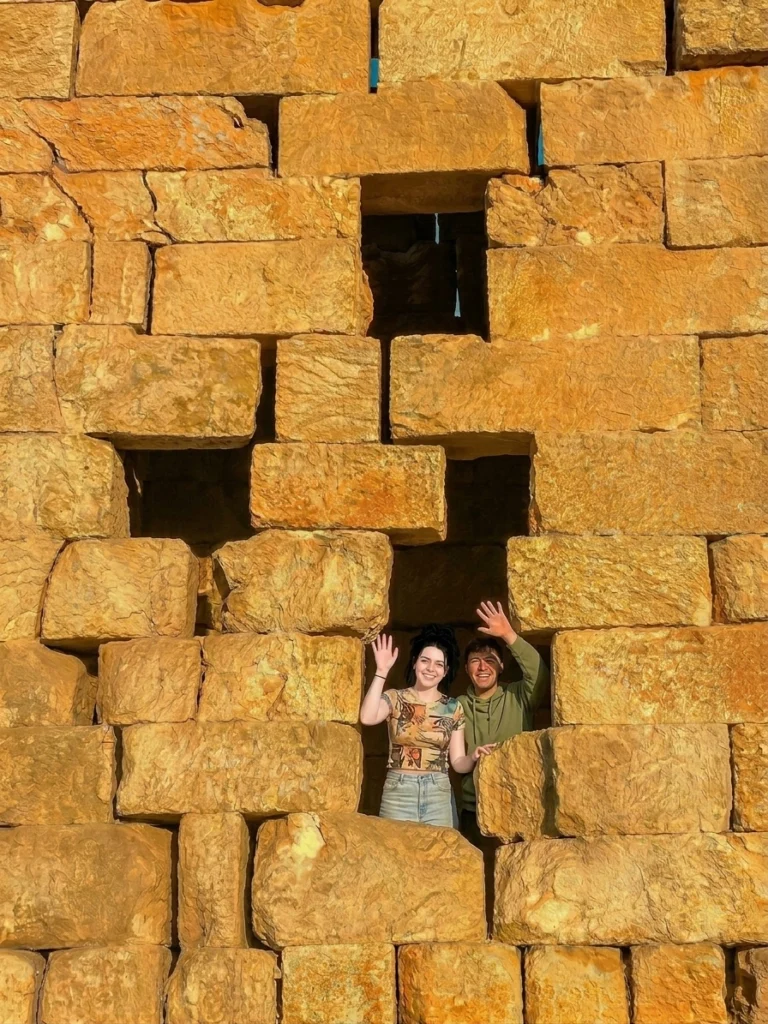 A high-angle, close-up photograph of a massive, golden-yellow stone wall featuring unique interlocking ashlar masonry at an ancient Byzantine site in the Dead Cities of Syria. Two young tourists—a woman with dark hair in a patterned top and jeans, and a young man in a green hoodie—smile and wave at the camera from inside a geometric, keyhole-style stone window cut into the historic facade.
