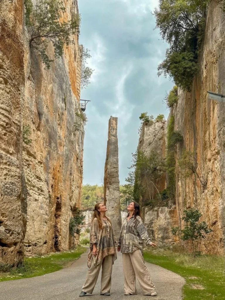 A dramatic, vertical low-angle shot of the monumental rock-cut moat at the UNESCO-listed Saladin Citadel (Sahyon Castle) near Latakia, Syria. Two Russian female tourists in wide, comfortable patterned tunic blouses and light beige trousers stand on the paved access road, looking up at the towering, sheer rock canyon walls under an overcast sky. In the center background stands the famous tall, slender limestone pillar left by medieval stonecutters to support the drawbridge.