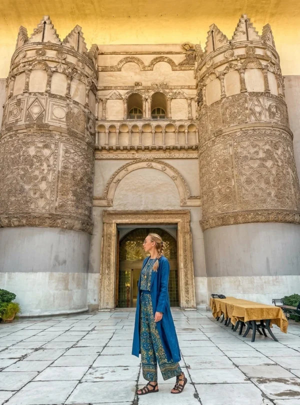 A blonde woman in a matching blue and patterned outfit stands on a paved courtyard, looking at the monumental gateway façade of the National Museum of Aleppo, Syria, which is formed by the reconstructed Qasr al-Hayr al-Gharbi palace gate.