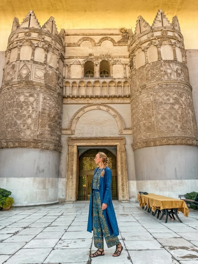 A blonde woman in a matching blue and patterned outfit stands on a paved courtyard, looking at the monumental gateway façade of the National Museum of Aleppo, Syria, which is formed by the reconstructed Qasr al-Hayr al-Gharbi palace gate.