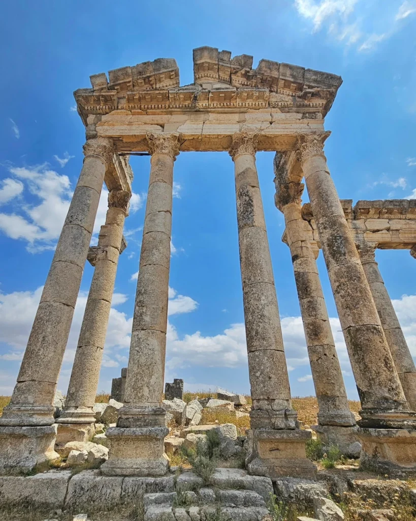 A low-angle view looking up at the towering stone Corinthian columns and carved entablature of the ancient colonnade at Apamea, Syria, set against a bright blue sky with light clouds.