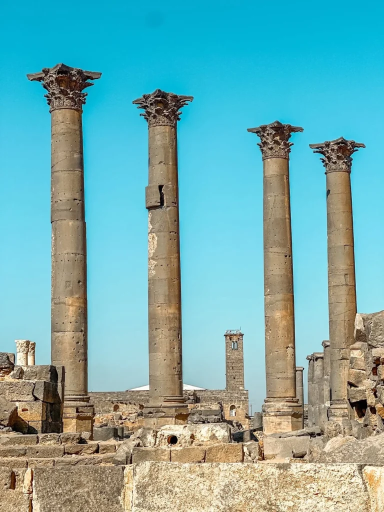 Four tall ancient black basalt Corinthian columns rising above stone ruins in Bosra, Syria, framed against a vibrant blue sky with historic stone structures in the distance.