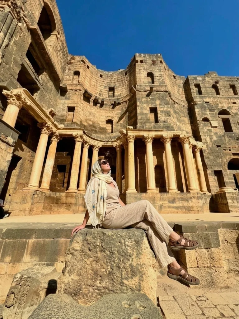 A European traveler in comfortable, neutral-toned linen clothing and sunglasses sitting on an ancient basalt stone block, gazing up at the grand stage facade and classical columns of the Roman Theatre of Bosra under a clear blue sky.
