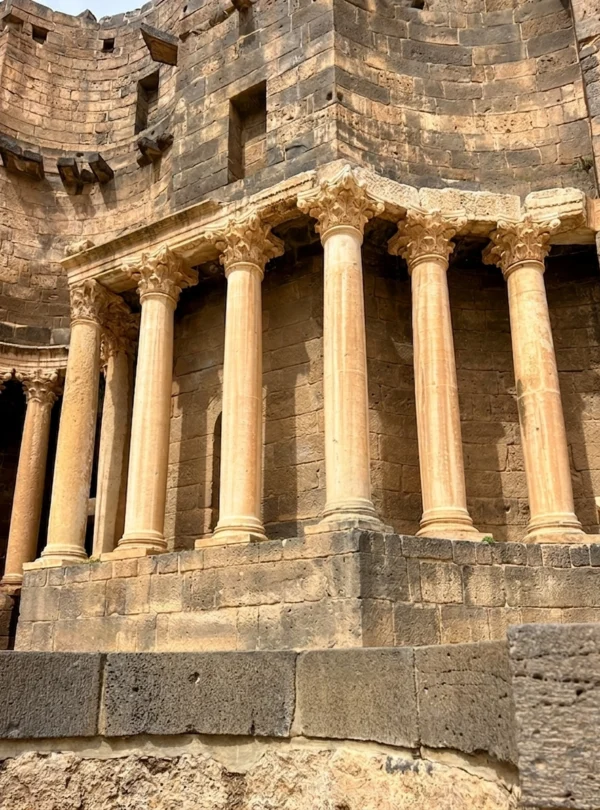 A detail view of the preserved Roman Theatre in Busra Sham, showing five ornate Corinthian columns supporting a lintel against a massive, curved wall of dark ancient basalt stone.
