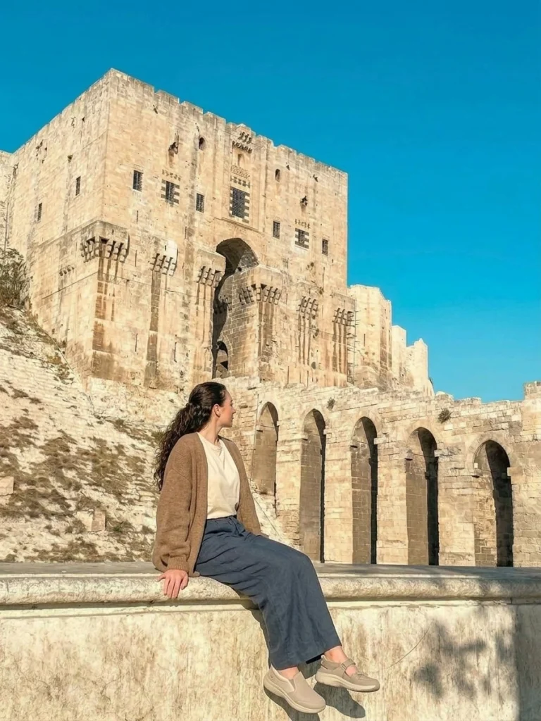 A European tourist seated on a stone ledge wearing comfortable neutral-toned travel attire, gazing toward the towering stone entrance gateway and arched bridge of the Citadel of Aleppo under a clear blue sky.