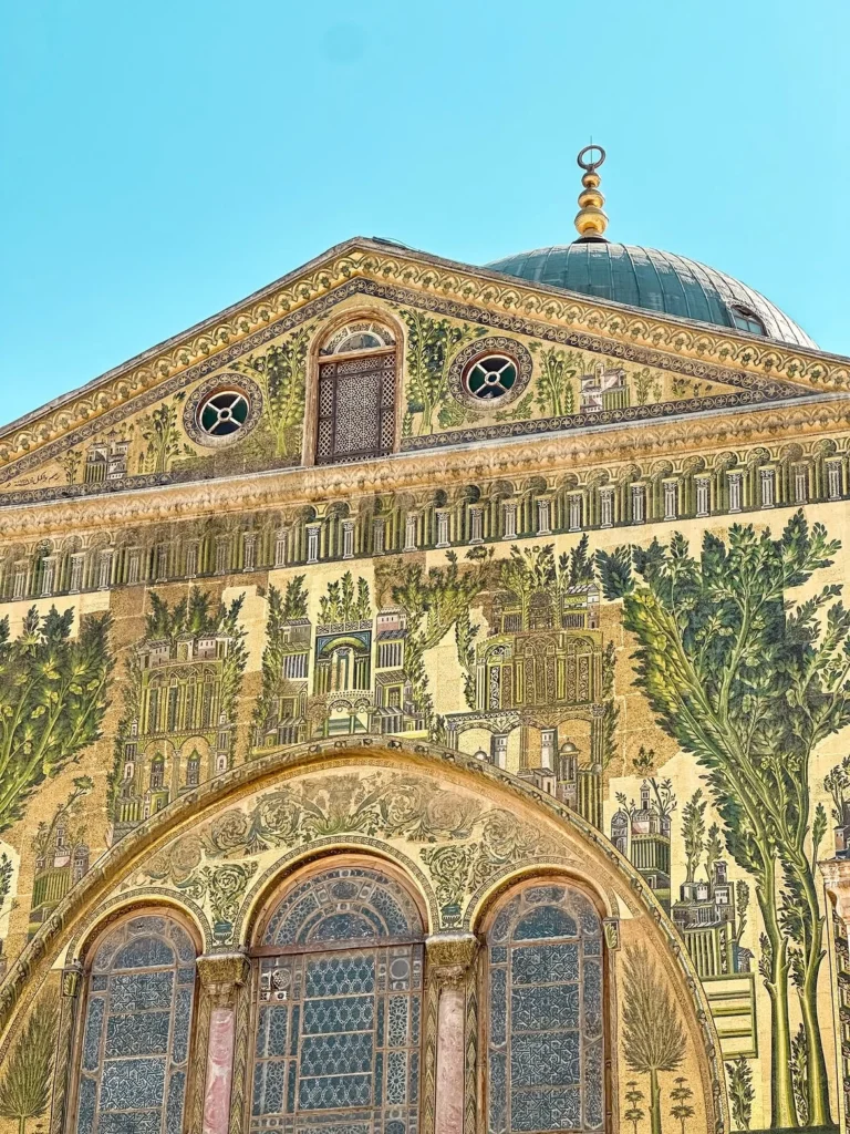 A detailed close-up of the courtyard façade of the Umayyad Mosque in Damascus, showing intricate golden and green wall mosaics depicting paradise and landscape scenes, set against marble arches and a copper-domed structure under a clear sky.