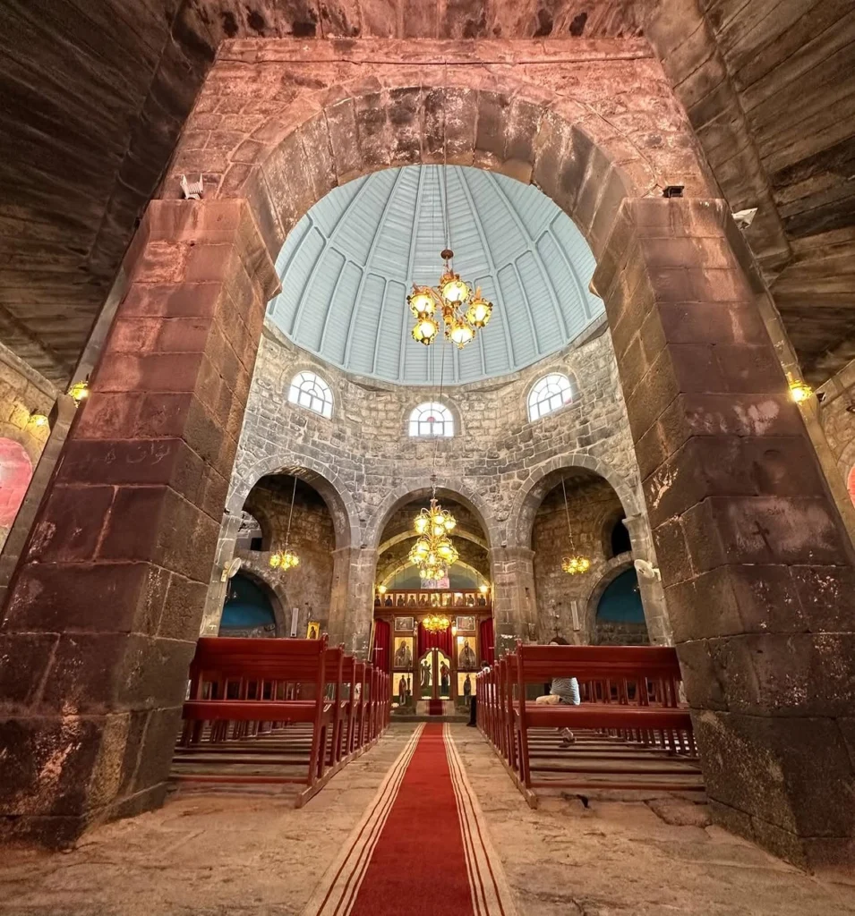 Interior perspective of Saint George Basilica in Izra’a, showing dark basalt stone arches, wooden pews lining a red aisle carpet, hanging chandeliers, and an octave dome ceiling.