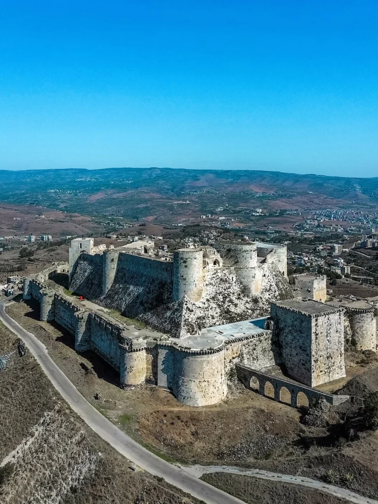 An elevated aerial view of the Krak des Chevaliers fortress in Syria, highlighting its massive stone ramparts, outer curtain walls, inner citadel, and aqueduct set against rolling hills under a clear blue sky.