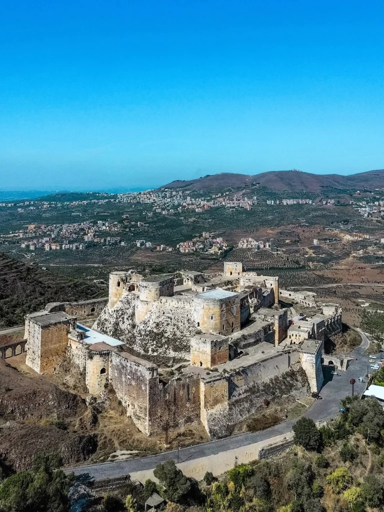 An expansive aerial view of the medieval Krak des Chevaliers fortress with its concentric defensive walls, stone towers, and central citadel set against rolling green hills and distant mountain villages under a clear blue sky.
