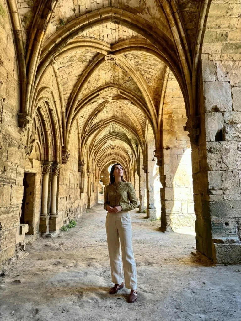 A traveler standing beneath the stone vaulted arches of the Gothic Hall loggia inside Krak des Chevaliers in Syria, looking up at the historic stonework bathed in warm natural light.