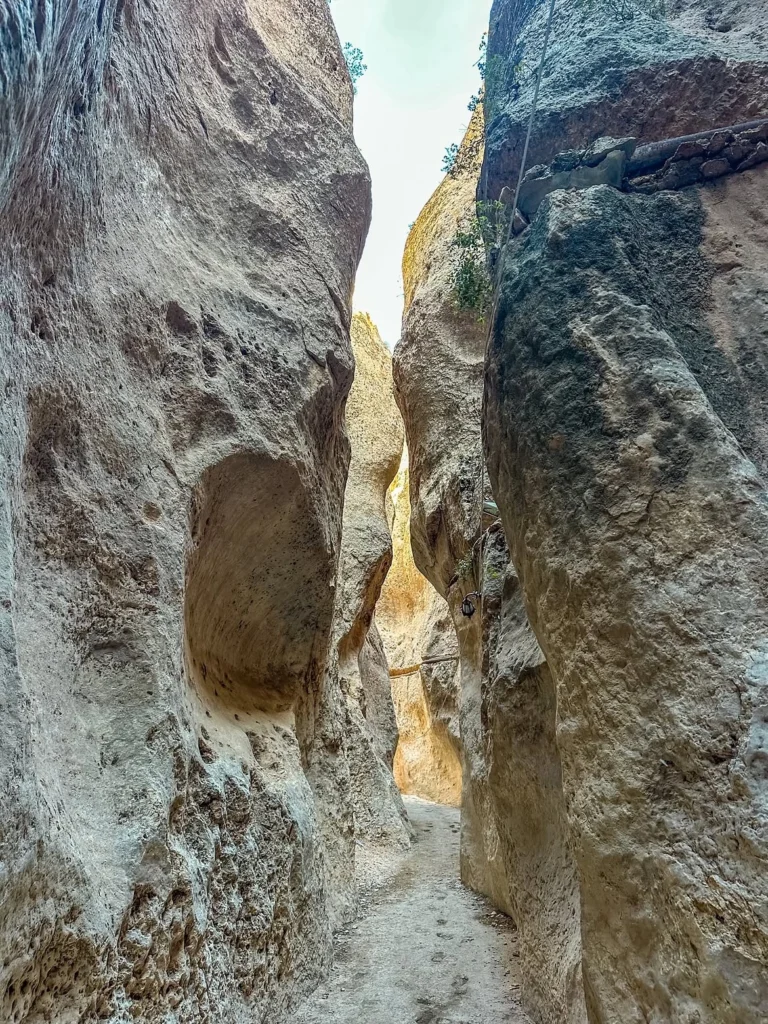 A narrow view from inside the historical Maaloula canyon in Syria, with a dirt path winding between towering, textured rock formations. The walls show intricate erosion and some embedded historical pipes, leading towards a sunlit opening at the far end of the passage.