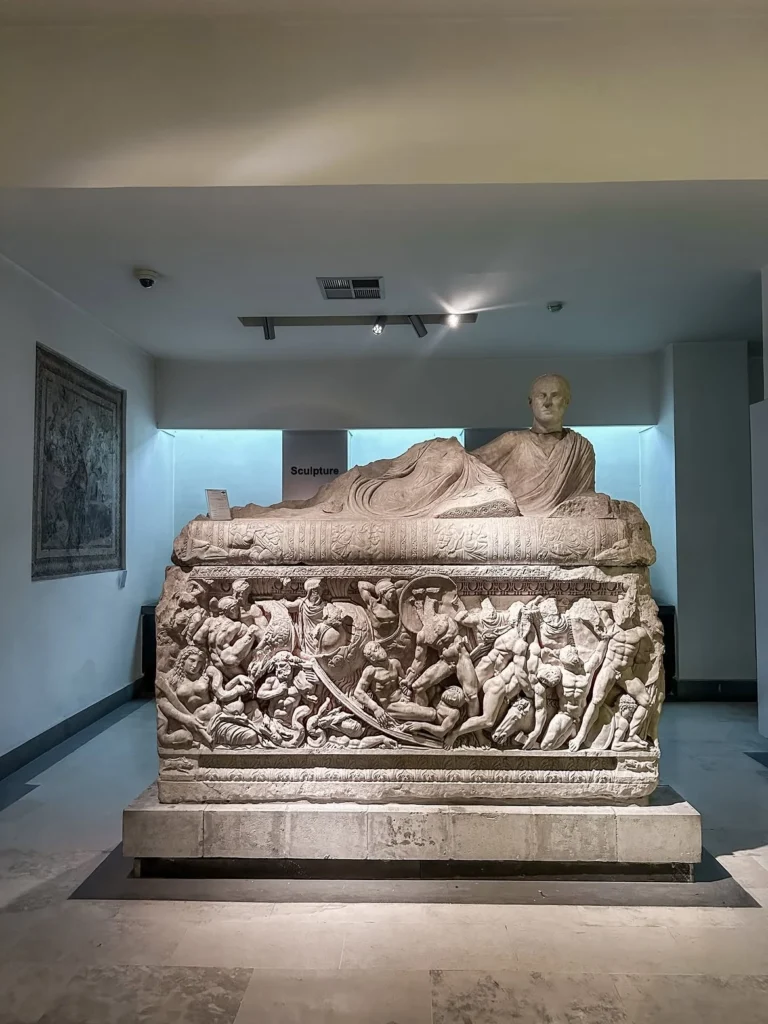 An intricately carved Roman marble sarcophagus featuring high-relief mythological battle figures along its base and a reclining noble statue on the lid, exhibited inside the National Museum of Damascus.