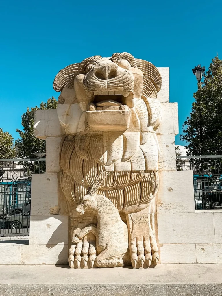 The monumental stone sculpture of the Lion of Al-lat protecting a kneeling gazelle between its paws, displayed outside the National Museum of Damascus against a clear blue sky.