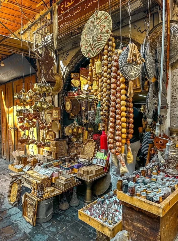 A vibrant craft stall in the covered souks of Old Damascus displaying long strings of oversized wooden prayer beads, hand-engraved brass coffee pots, inlaid wooden boxes, and traditional spices under vaulted stone alleyways.