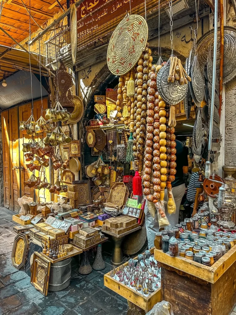 A vibrant craft stall in the covered souks of Old Damascus displaying long strings of oversized wooden prayer beads, hand-engraved brass coffee pots, inlaid wooden boxes, and traditional spices under vaulted stone alleyways.