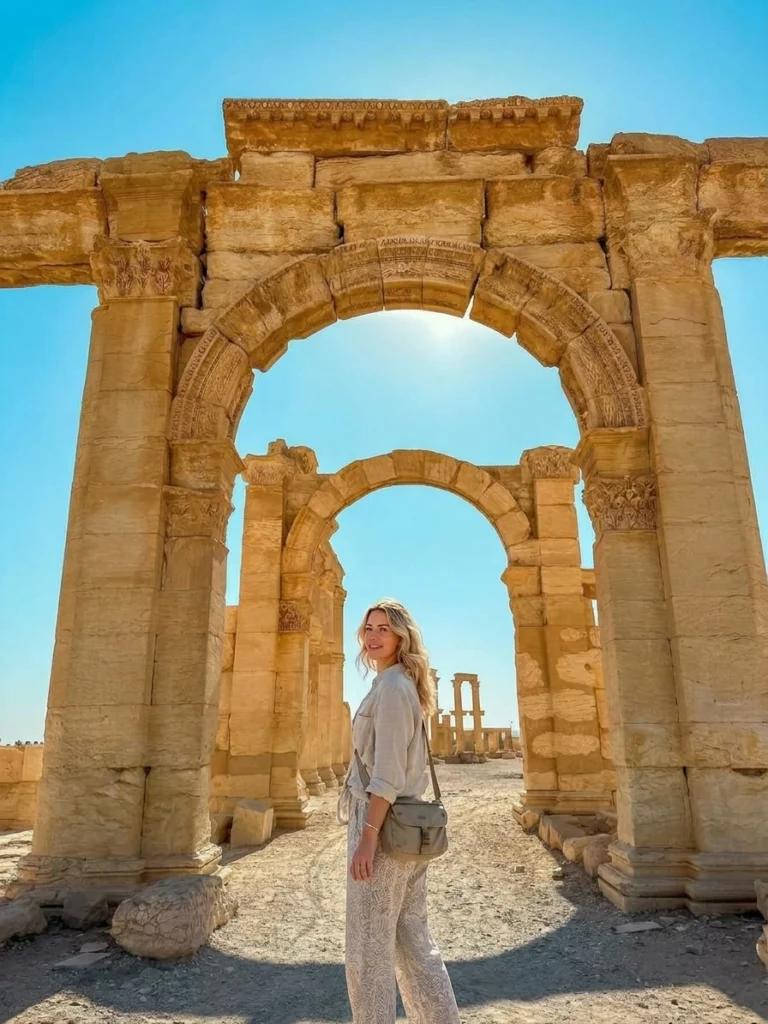 A blonde traveler standing under the ancient stone arches of Palmyra, Syria, looking back over her shoulder, with sandy terrain and towering Roman-era colonnades stretching into the background beneath a clear blue sky.