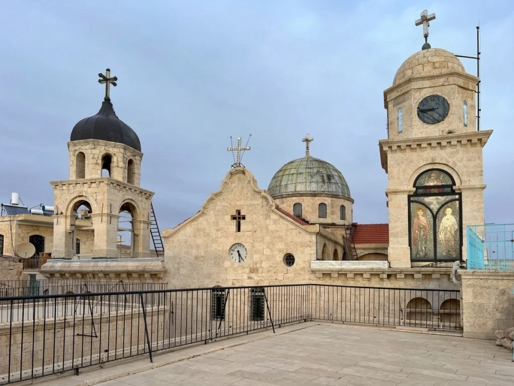 A view of the upper courtyard and rooftop terrace at the Convent of Our Lady of Saidnaya in Syria, showcasing its historic stone bell towers, crosses, tiled domes, and mosaic artwork set against an overcast sky.
